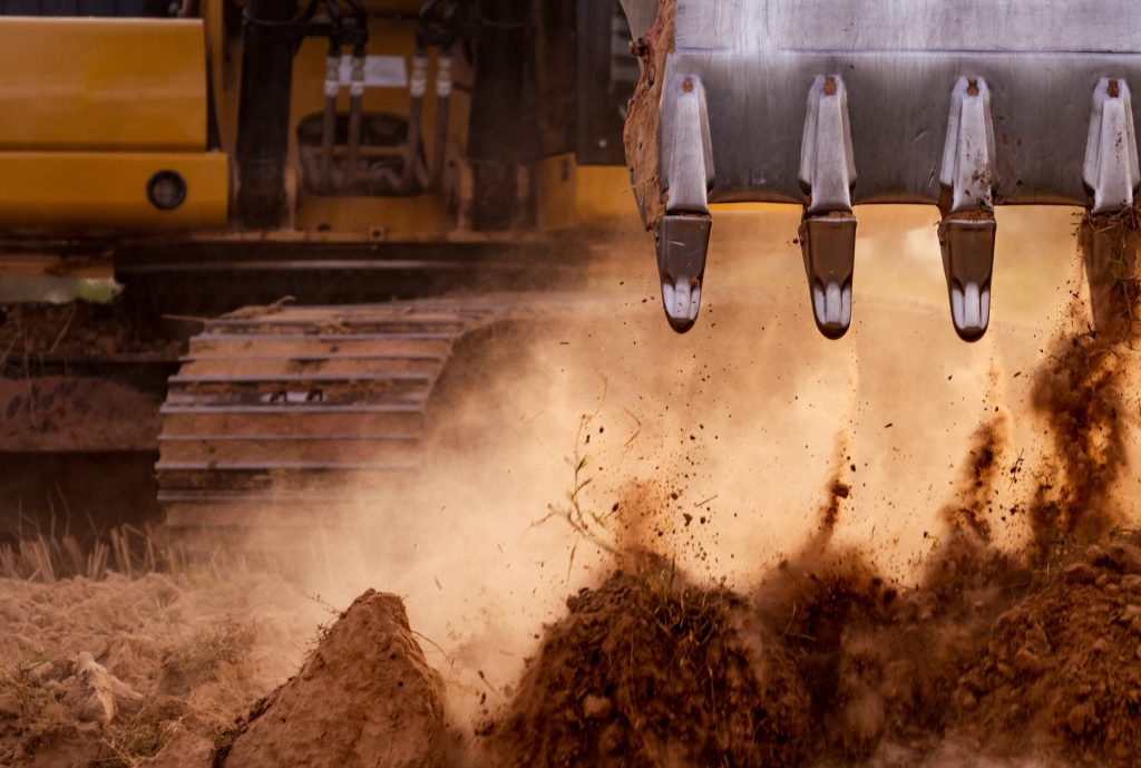 Selective focus on metal bucket teeth of backhoe digging soil. Backhoe working by digging soil at construction site. Crawler excavator digging on soil. Earth moving machine. Excavation vehicle.