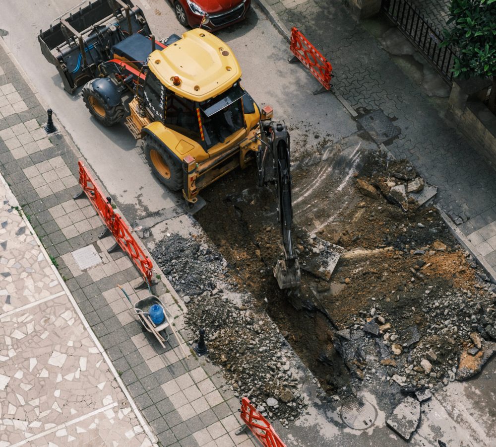 An excavator repairs sewer storm hatches ata street on a summer